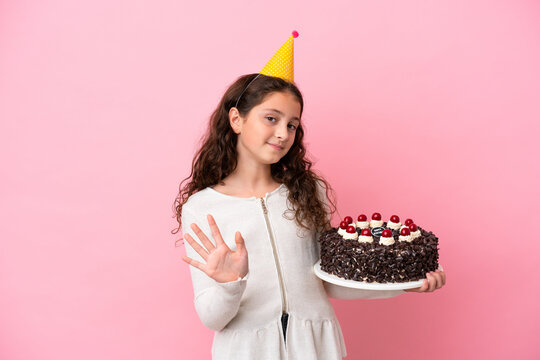 Little caucasian girl holding birthday cake isolated on pink background saluting with hand with happy expression