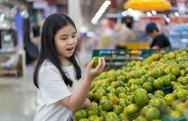 Asian child holding sweet green orange in pile or kid girl happy smile to like eat fruit for vitamin c and wearing white t shirt to shopping and buy in super market and department store