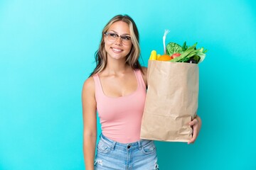 Young Russian woman holding a grocery shopping bag isolated on blue background looking to the side and smiling