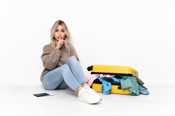 Teenager blonde girl with a suitcase full of clothes sitting on the floor showing a sign of silence gesture putting finger in mouth