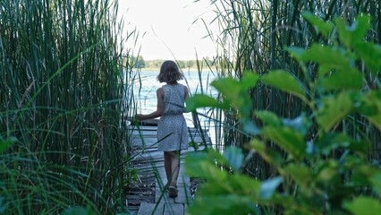 Cute Young Caucasian Girl Walking on Old Wooden Pier Through Reed Bed Rushes at Serene Lake Shore