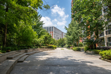 Public outdoor area with pavement tiles, surrounded by urban buildings and lush green trees providing shade, concrete garden beds with shrubs. A landscaped public garden in a Chinese neighbourhood