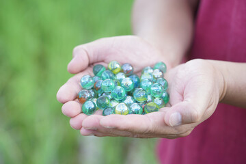 Close up hands hold round small marble balls, spherical objects often made from glass. Green background. Concept, object use as toy for playing games or use for DIY craft decoration.     