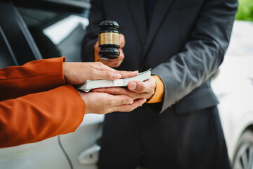 Two Asian individuals discuss a car insurance claim with an agent in front of a damaged vehicle. They review documents, discuss the policy, and address the crash and repair costs.