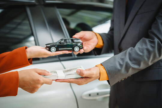 Two Asian individuals discuss a car insurance claim with an agent in front of a damaged vehicle. They review documents, discuss the policy, and address the crash and repair costs.