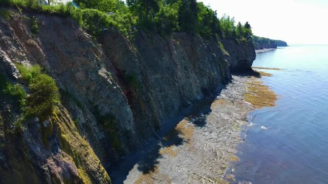 Un drone longe une falaise rocheuse qui s'&eacute;tend a perte de vue, coiff&eacute;e de v&eacute;g&eacute;tation sur le bord de la plage et de la mer tranquille de la Baie-des-Chaleurs, Gasp&eacute;sie, Qu&eacute;bec, Canada.
