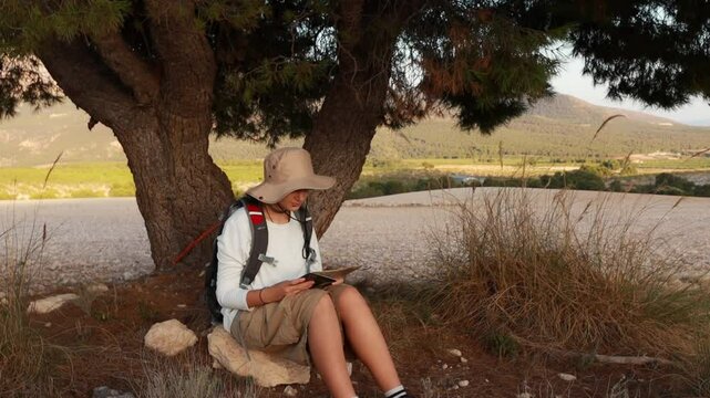 Hiker sits under tree, consulting map while enjoys hade. Girl Wearing hat and backpack,  hiker takes moment to plan  next adventure in serene outdoor setting