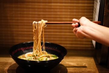 Crop anonymous female with chopsticks eating yummy ramen soup with noodles and meat in black bowl at ramen restaurant.