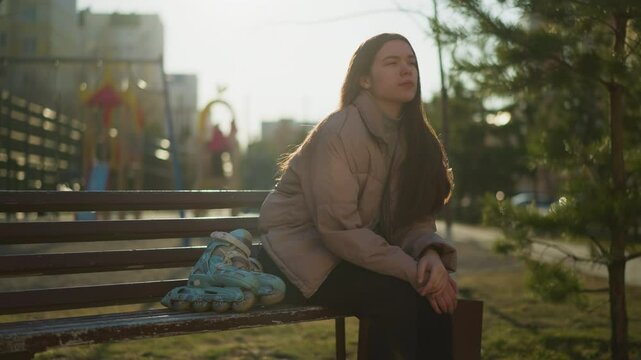 Young girl wearing a peach jacket, grey inner shirt, and black trousers sits on a park bench with her rollerblades beside her. She rests one hand on her chin in deep thought, with a blurred background