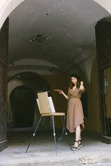 Girl in dress and black hat painting on an easel in a city on a street