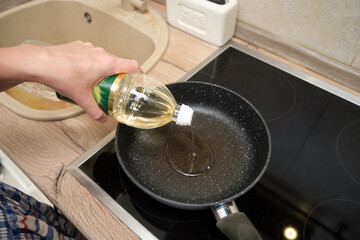 Hand pouring cooking oil into a black frying pan on an electric stove. Close-up kitchen scene showing food preparation