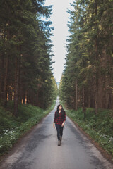 Fototapeta premium Young woman in a red plaid shirt walking down a peaceful, tree-lined forest road, embracing the tranquility and beauty of nature, symbolizing freedom and a journey of self-discovery