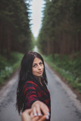 Naklejka premium Young woman with long dark hair in a red checkered shirt, standing on a forest road, holding hand while looking back. The image captures a moment of connection in nature