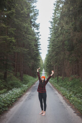 Smiling woman in a red checkered shirt and bright orange beanie stands in the middle of a forest road, stretching her arms behind her head, enjoying the tranquility of the surroundings