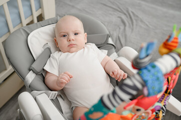 Serious little baby comfortably settled in baby rocking chair surrounded by colorful safe baby development toys. Baby is entertained during waking hours