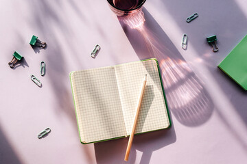 Modern summer stationery still life. A glass of water with long shadow and open notepad with pencil on purple background. Top view, flat lay
