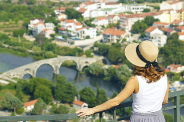 Fototapeta premium Photographed from behind, a young woman in a hat admires the view from above of the main landmark of the city of Trebinje - the Arslanagic (Perovic) Bridge. A female tourist in Bosnia and Herzegovina.
