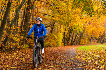 Middle aged woman wearing blue sporty jacket, black cycling pants and white bike helmet riding bicycle in city forest in autumn scenery. Front view.