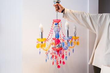 An anonymous hand holds a retro and vintage style chandelier lamp on a plain white background. The lamp is made of crystals of many colors with arms and hanging rhinestones.