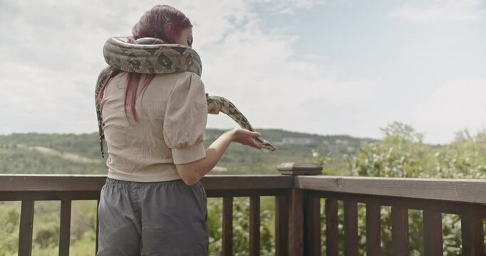 A woman with red hair holds her pet snake on a balcony overlooking a beautiful vista