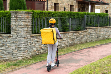 Latin food delivery girl with yellow backpack riding an electric scooter in a luxury neighborhood