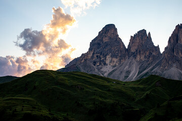 Majestic mountain peaks under a colorful sunset with rolling green hills in the foreground and clouds painting the evening sky