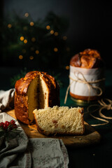 Two homemade Christmas panettone , one cut and the other whole, made as holiday present with pine and red berries decorations with Christmas lights on green background.