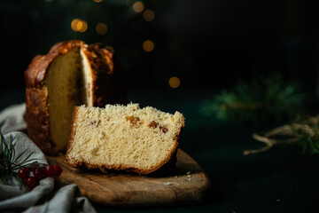 Sliced homemade Christmas panettone, made as holiday present with pine and red berries decorations on wooden table on rustic kitchen with Christmas lights on background.