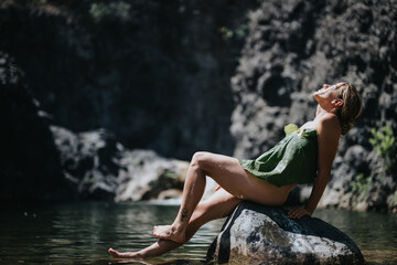 A young woman in a leaf dress relaxing on a rock by the river, enjoying nature and the sunlight.