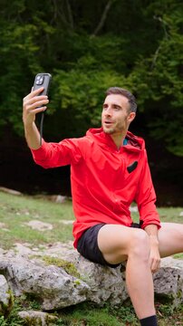 Fitness coach live streaming on phone in nature. Young man talking on a video call in a forest. Positive influencer creating content for social media outdoors.