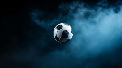 Fototapeta premium A soccer ball is suspended in mid-air against a dark, smoky background. The ball is illuminated, showcasing its texture and detail. The smoke adds a sense of motion and intensity to the image, suggest