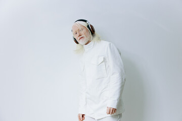 Elderly man with long white hair and headphones wearing white clothing against a light gray...