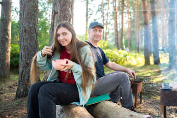 Teenage girl eating chocolate balls with milk for breakfast, sitting next to her dad, at a picnic, in a tent camp in a pine forest. focus on the girl