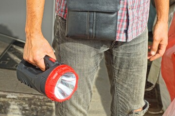 one big round electric powerful plastic black with white reflector modern new flashlight in hand of man dressed in gray jeans and checkered shirt with leather small bag daytime outdoors