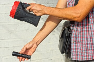 two big and small round electric powerful plastic black with white reflector modern new flashlights in the hands of a man dressed in gray jeans and a checkered shirt with a leather small bag 