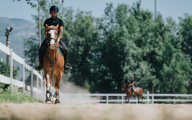 Man riding horse at equestrian facility on a sunny day. The scene features white fences and lush green trees in the background.