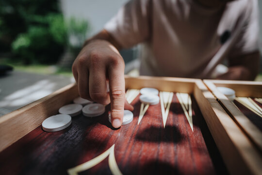 Close-up image of a person playing backgammon outdoors, capturing the strategic movement of pieces on a wooden board under bright sunlight.