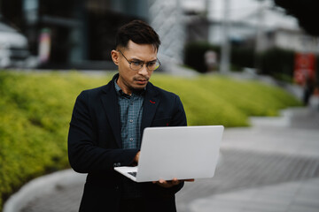 Focused Professional: A young man in a business suit and glasses intently works on his laptop, his concentrated expression reflecting dedication and ambition. The modern urban backdrop adds to the sen © Wicitr
