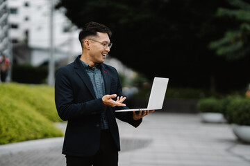 Businessman on the Go: A confident Asian businessman in a sleek suit makes a video call on his laptop outdoors, embodying the modern, mobile professional.  