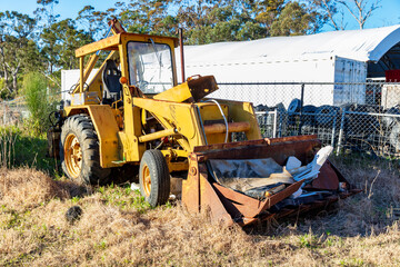 Photograph of an old and rusty industrial front end loader with a bucket full of rubbish that has been abandoned amongst other trash in a grassy field.