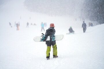  Panoramic view of a ski slope with a woman going up the slope with a snowboard behind her in the foreground. It's snowing.