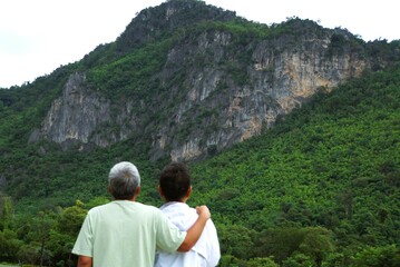 Life partner Asian middle-aged couple standing with arms around shoulders looking at large mountains view. Feeling warmth, comfort  happiness in National park Thailand, Living happy life retirement