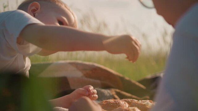 A close-up shot of two young boys sitting in a grassy field, sharing bread. The younger boy tenderly offers a small piece of bread to his older brother, capturing a moment of sibling bonding and care