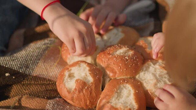 Close-up shot of children's hands pinching and eating freshly baked sesame bread rolls on a checkered picnic blanket. The scene captures a simple and peaceful outdoor moment