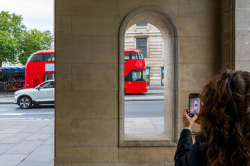 Capturing London’s Iconic red buses: A tourist’s snapshot through historic arches on a bustling city street in the UK