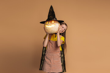 Smiling little girl wearing witch costume holding jack-o'-lantern, isolated on beige background