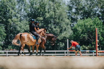 Two individuals are horseback riding in an outdoor equestrian training session while a coach adjusts obstacles in the foreground.