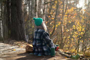 Rearview of serene woman backpacker meditating on eco trail, watching natural landscape in sunny fall woodland. Calm female enjoy autumn air, basking in sun, drinking hot thermos tea in park