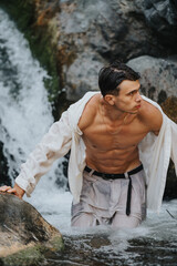 Young man emerging from waterfall in rocky terrain, displaying fitness and adventure in nature, with focus on his toned body and determined expression.