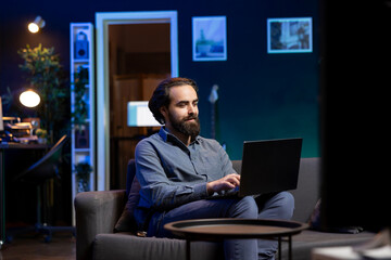 Smiling freelancer working on laptop while watching live sports event on TV in living room. Remote worker at home typing on notebook, solving business tasks and keeping football match in background